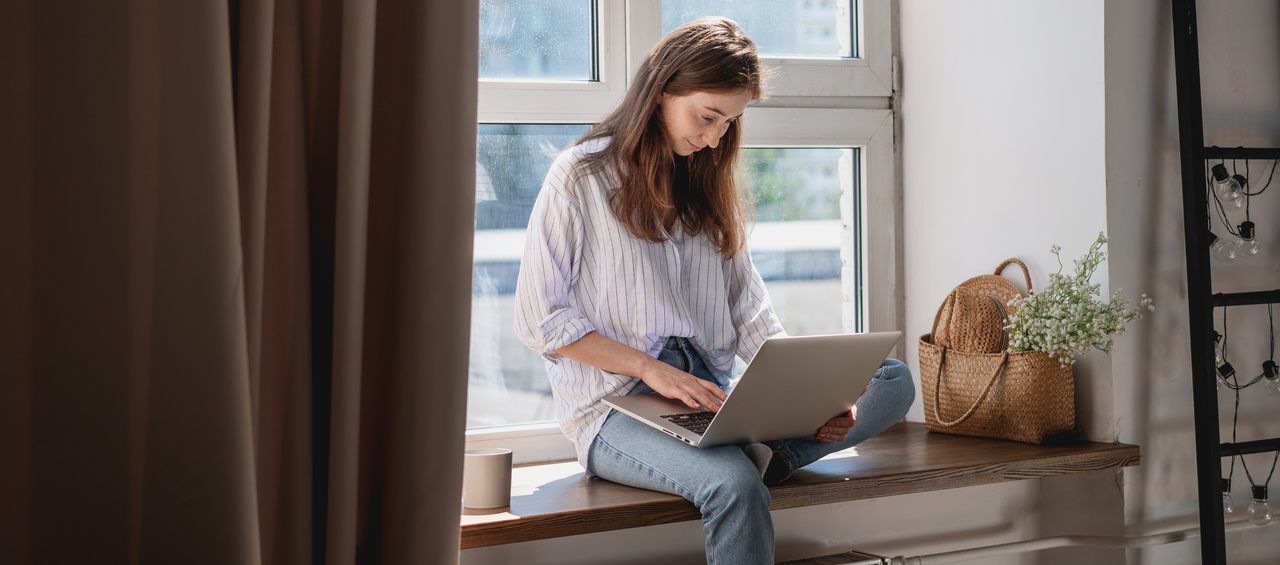 a woman sitting in front of the window and looking at her laptop screen and smiling